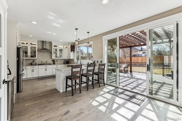 kitchen featuring wall chimney exhaust hood, backsplash, appliances with stainless steel finishes, glass insert cabinets, and a kitchen breakfast bar