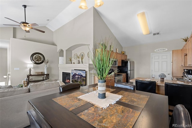 dining area with ceiling fan, arched walkways, high vaulted ceiling, and a glass covered fireplace