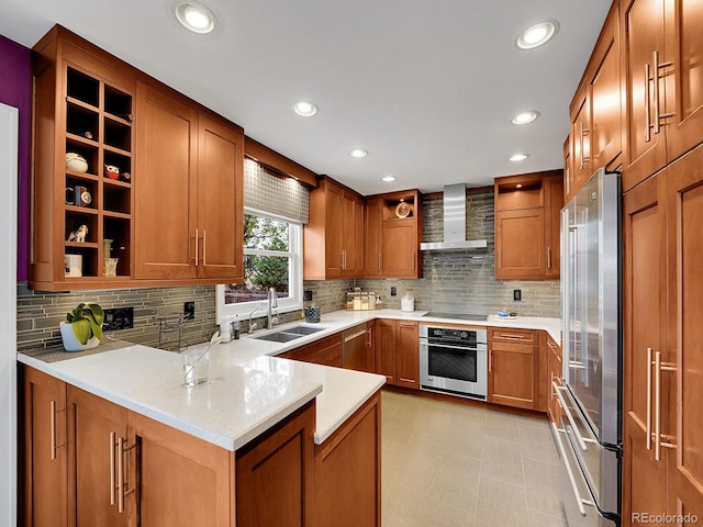 kitchen featuring brown cabinetry, a peninsula, wall chimney range hood, a sink, and stainless steel appliances