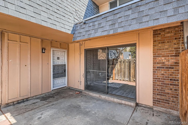 doorway to property with mansard roof, roof with shingles, a patio, and brick siding