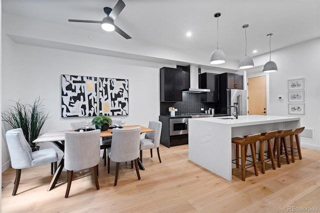 kitchen featuring pendant lighting, wall chimney range hood, appliances with stainless steel finishes, tasteful backsplash, and an island with sink