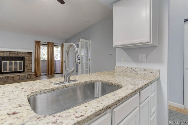 kitchen featuring ceiling fan, a stone fireplace, light stone counters, a sink, and white cabinetry