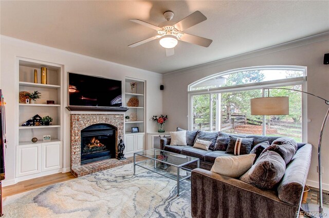 living room with crown molding, a brick fireplace, a ceiling fan, built in shelves, and wood finished floors
