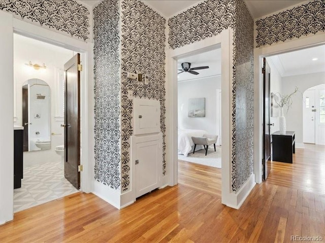 corridor with arched walkways, light wood-type flooring, wallpapered walls, ornamental molding, and baseboards