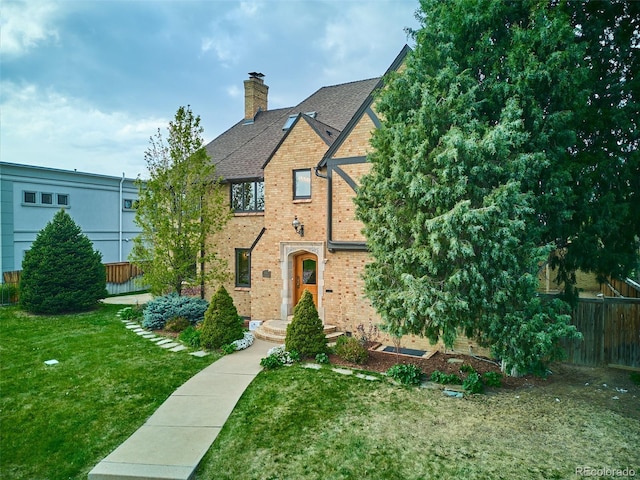 english style home with a chimney, brick siding, fence, a shingled roof, and a front lawn