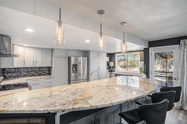 kitchen with light wood finished floors, backsplash, wall chimney range hood, light stone counters, and stainless steel refrigerator with ice dispenser