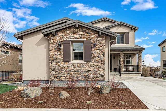 view of front facade with covered porch, a tile roof, fence, stone siding, and stucco siding