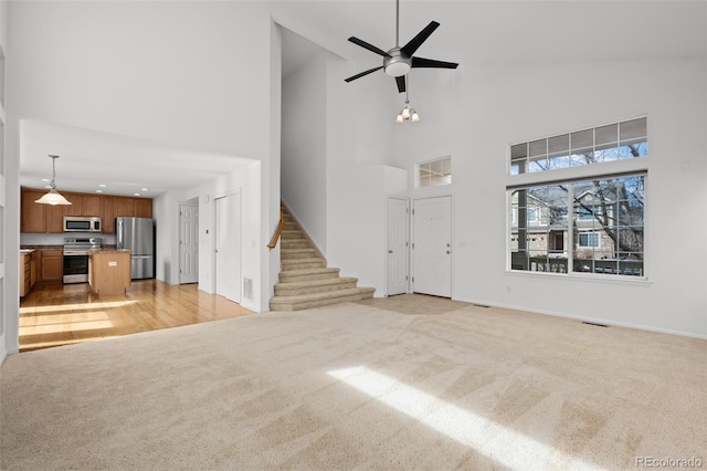 unfurnished living room featuring high vaulted ceiling, light colored carpet, ceiling fan, and stairs