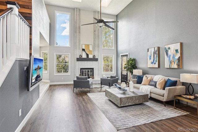 living room with ceiling fan, a brick fireplace, baseboards, wood finished floors, and a towering ceiling