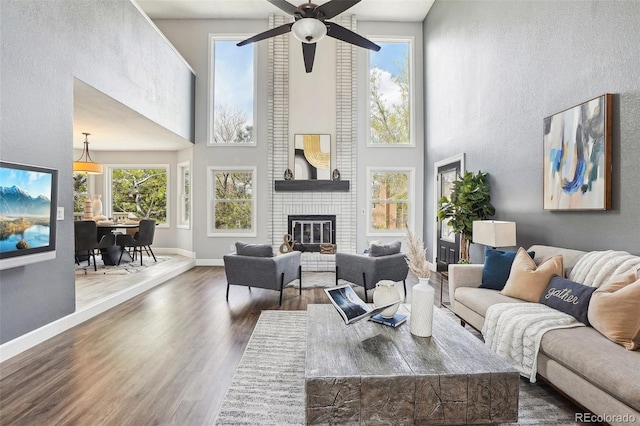 living room featuring a fireplace, a ceiling fan, baseboards, wood finished floors, and a towering ceiling