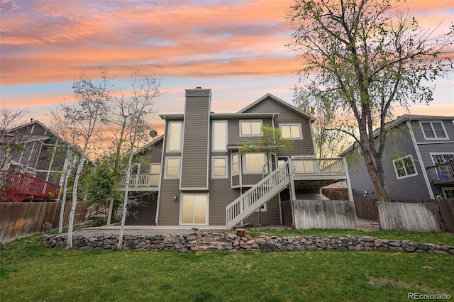 back of house at dusk with fence, a deck, a chimney, a yard, and stairway