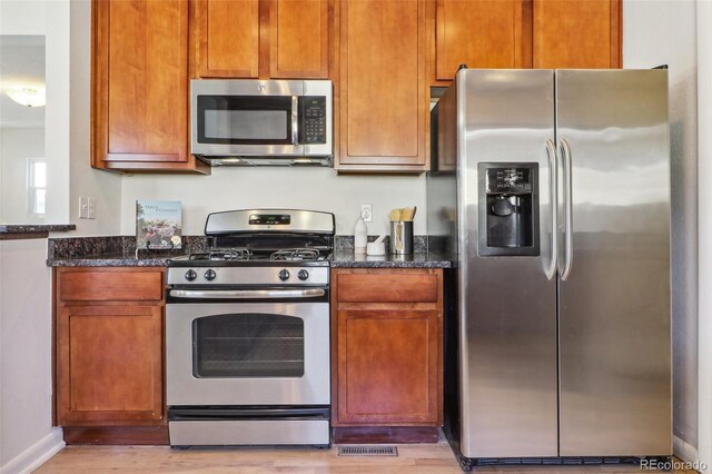 kitchen featuring light wood-type flooring, brown cabinetry, dark stone countertops, and appliances with stainless steel finishes