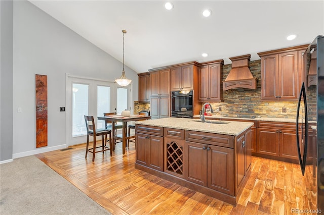 kitchen with pendant lighting, premium range hood, black appliances, a center island with sink, and light wood-type flooring