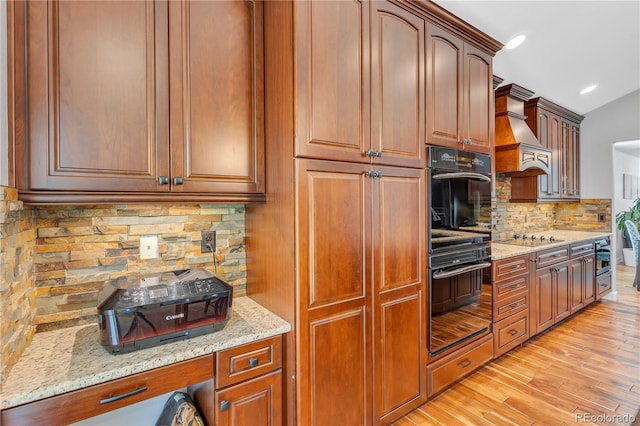 kitchen with light stone counters, backsplash, light wood-type flooring, black appliances, and custom exhaust hood