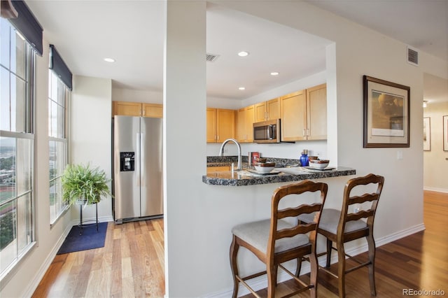 kitchen with a kitchen bar, appliances with stainless steel finishes, light brown cabinetry, a healthy amount of sunlight, and light hardwood / wood-style flooring