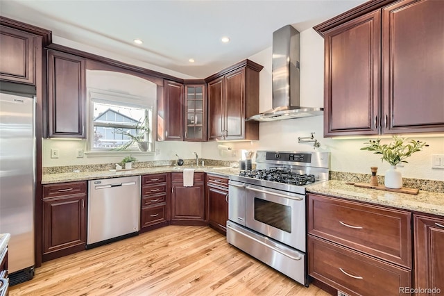 kitchen featuring light stone counters, appliances with stainless steel finishes, wall chimney exhaust hood, and light wood-type flooring