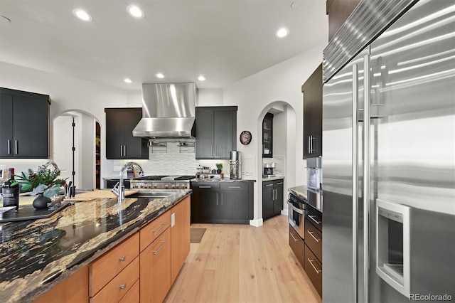 1287 Buffalo Ridge Rd, Castle Pines, CO 80108 - kitchen with stainless steel appliances, arched walkways, wall chimney range hood, light wood-style floors, and recessed lighting