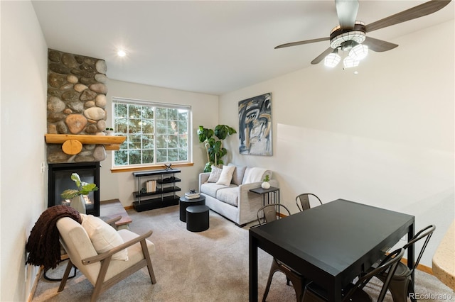 living area featuring a ceiling fan, carpet flooring, a stone fireplace, and baseboards