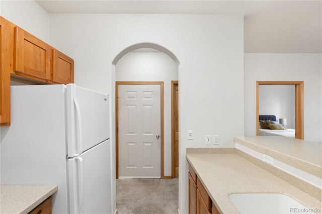 kitchen with arched walkways, light colored carpet, light countertops, freestanding refrigerator, and brown cabinetry