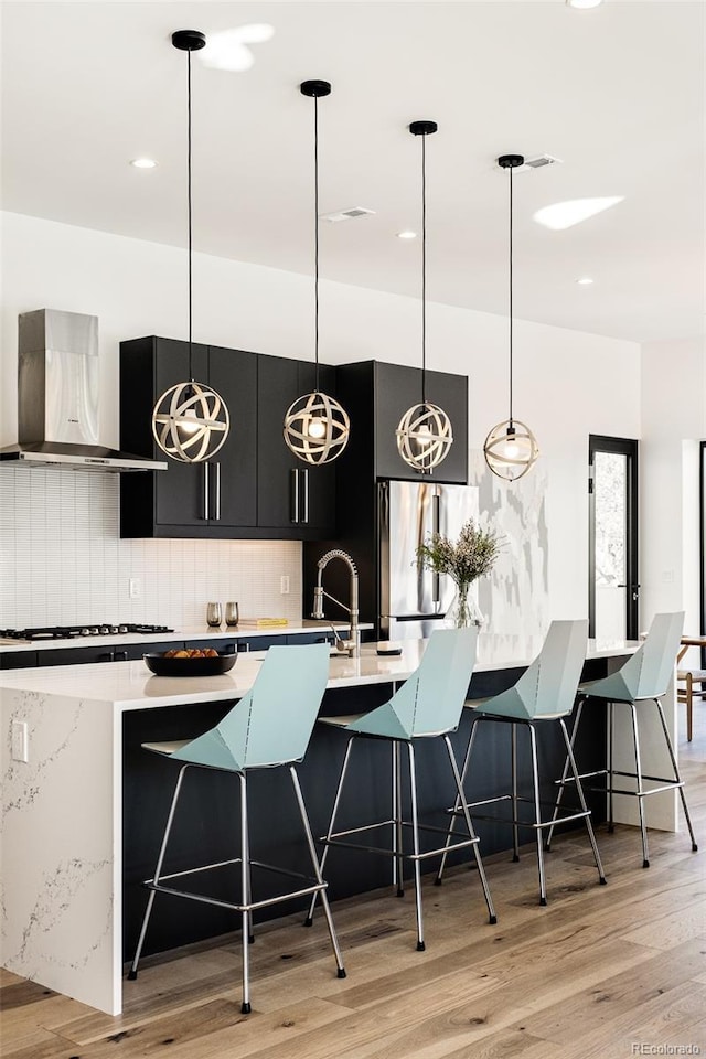 kitchen featuring stainless steel refrigerator, wall chimney range hood, decorative light fixtures, a breakfast bar area, and decorative backsplash