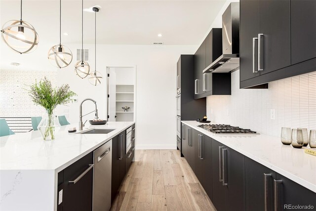 kitchen featuring light wood-type flooring, stainless steel appliances, sink, wall chimney range hood, and decorative light fixtures