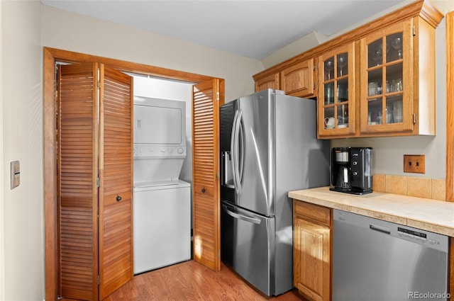 kitchen featuring brown cabinets, stainless steel appliances, stacked washer and dryer, light countertops, and glass insert cabinets