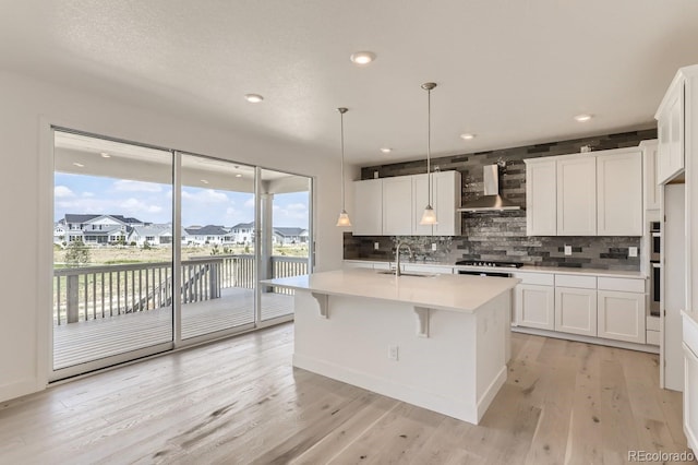 kitchen featuring a kitchen island with sink, wall chimney exhaust hood, light wood-type flooring, decorative light fixtures, and white cabinetry