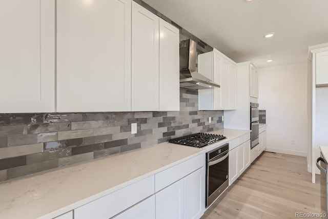 kitchen with white cabinetry, light wood-style floors, appliances with stainless steel finishes, wall chimney exhaust hood, and decorative backsplash