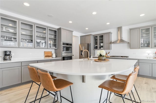 kitchen featuring wall chimney exhaust hood, gray cabinetry, stainless steel appliances, light wood-type flooring, and a center island with sink