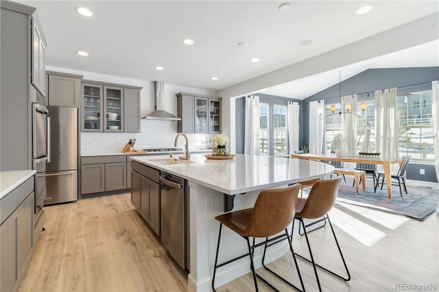 kitchen with wall chimney range hood, light hardwood / wood-style floors, a center island with sink, sink, and stainless steel appliances