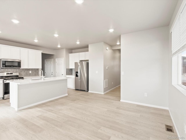 kitchen featuring white cabinetry, a center island with sink, appliances with stainless steel finishes, decorative backsplash, and light wood-type flooring