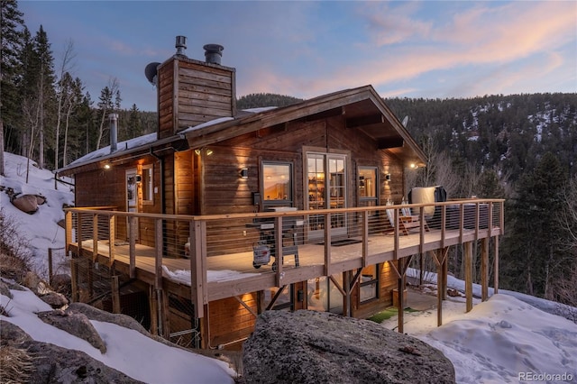 snow covered back of property featuring a chimney, a wooden deck, and a forest view