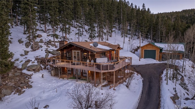 snow covered back of property with a deck, a chimney, a view of trees, an outbuilding, and a detached garage