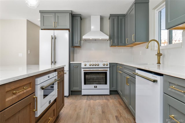 kitchen featuring light wood-type flooring, gray cabinets, a sink, white appliances, and wall chimney range hood