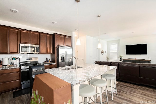 kitchen with sink, an island with sink, appliances with stainless steel finishes, decorative light fixtures, and light stone counters