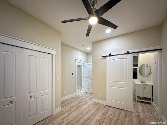 unfurnished bedroom with a closet, a barn door, light wood-style floors, a ceiling fan, and baseboards