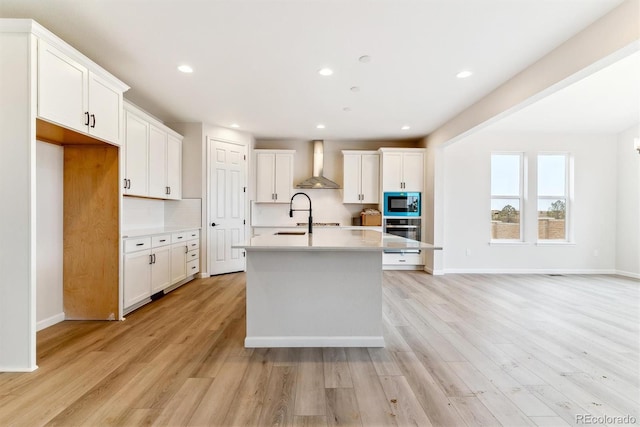 kitchen with white cabinets, wall chimney range hood, an island with sink, and appliances with stainless steel finishes