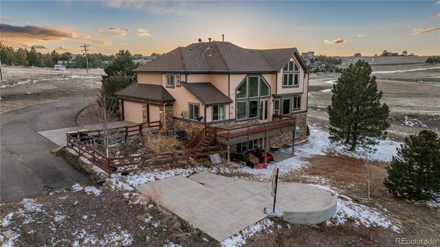 back of house at dusk with an attached garage, a shingled roof, a patio area, stone siding, and driveway