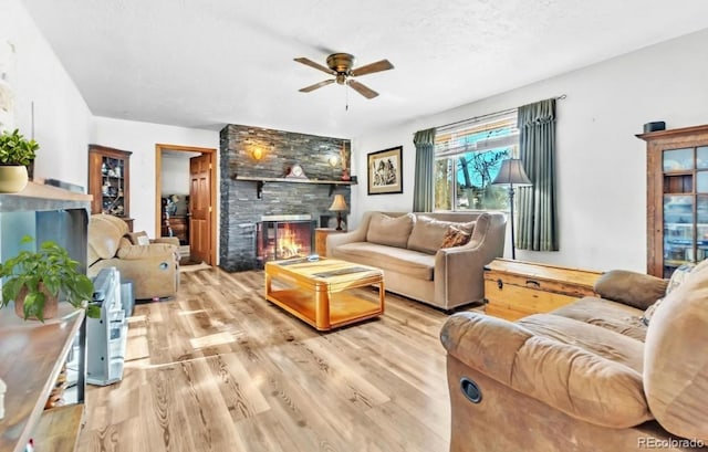 living room featuring ceiling fan, a stone fireplace, light hardwood / wood-style flooring, and a textured ceiling