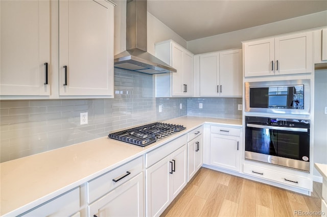 kitchen featuring tasteful backsplash, light wood-type flooring, wall chimney range hood, appliances with stainless steel finishes, and white cabinets