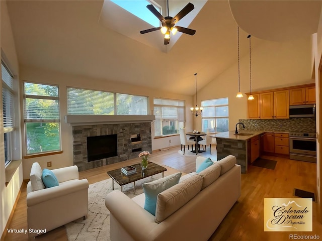 living area with ceiling fan with notable chandelier, high vaulted ceiling, light wood-type flooring, and a fireplace