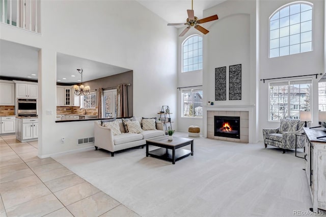 living room featuring visible vents, a tiled fireplace, ceiling fan with notable chandelier, light tile patterned flooring, and light colored carpet