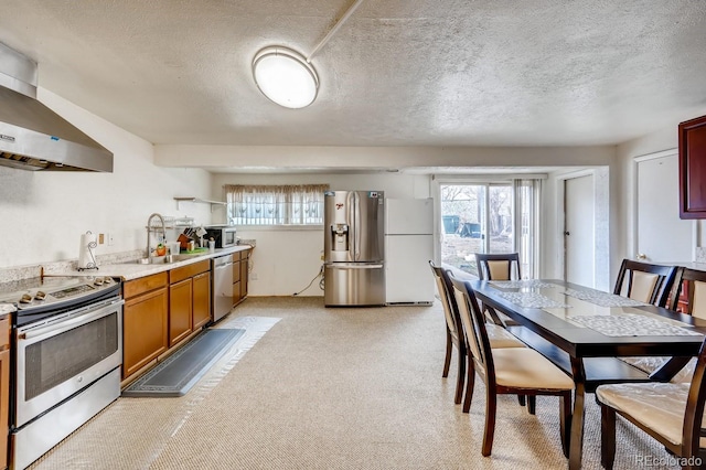 kitchen with wall chimney range hood, a sink, a textured ceiling, brown cabinets, and appliances with stainless steel finishes
