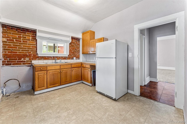 kitchen with brick wall, brown cabinets, freestanding refrigerator, vaulted ceiling, and a sink