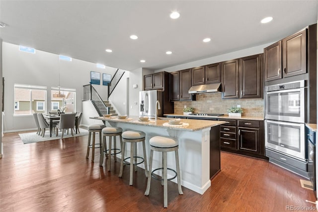 18180 W 84th Pl, Arvada, CO 80007 - kitchen featuring appliances with stainless steel finishes, under cabinet range hood, a breakfast bar, an island with sink, and dark brown cabinetry