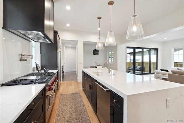 kitchen with appliances with stainless steel finishes, wall chimney range hood, sink, a large island with sink, and hanging light fixtures