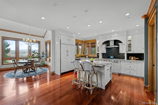 kitchen featuring wall chimney range hood, dark wood-style floors, a notable chandelier, a breakfast bar, and white cabinetry