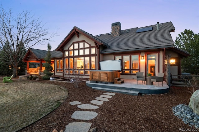 back of house at dusk featuring a wooden deck, a chimney, a tiled roof, a hot tub, and solar panels