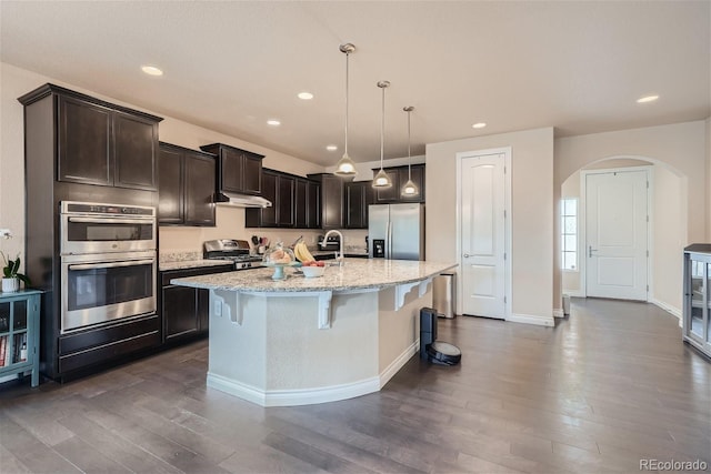 kitchen featuring stainless steel appliances, pendant lighting, an island with sink, light stone countertops, and dark hardwood / wood-style floors