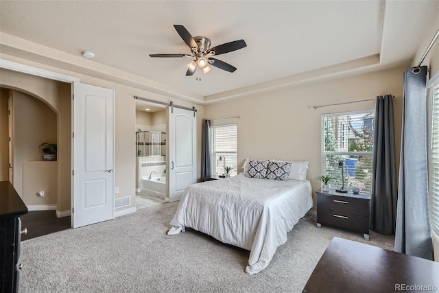 bedroom with a barn door, a tray ceiling, ceiling fan, carpet, and ensuite bath